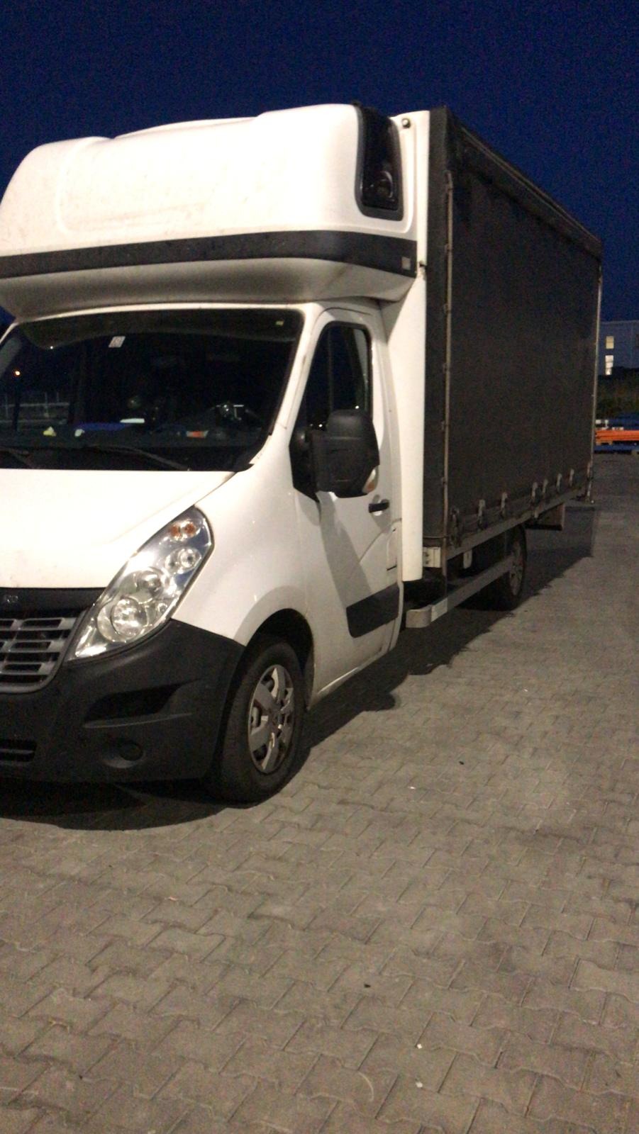 Biały samochód dostawczy z plandeką zaparkowany na brukowanym placu, sfotografowany z niskiej perspektywy przy słabym oświetleniu wieczornym.