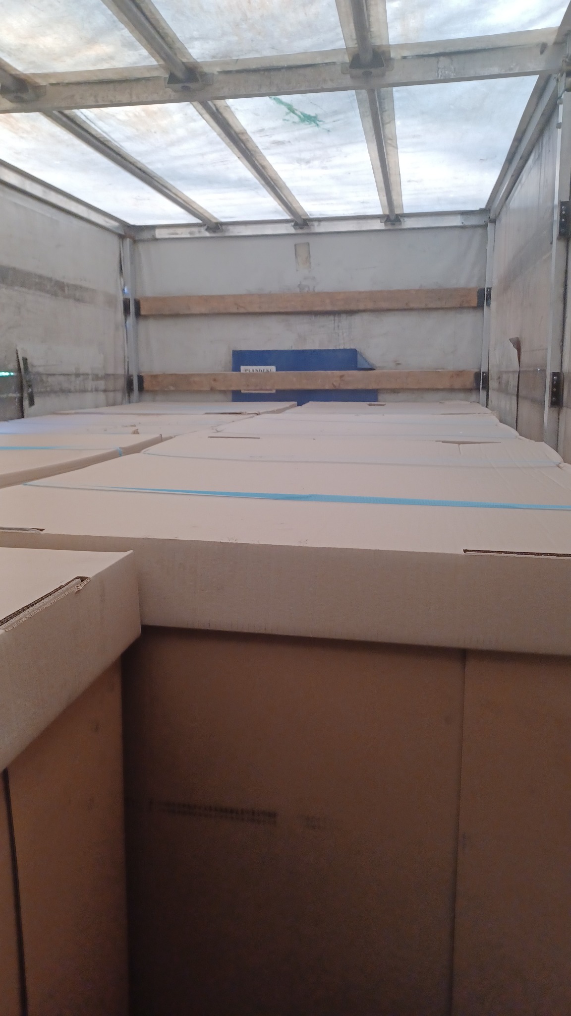 Wnętrze przestrzeni ładunkowej wypełnione po brzegi kartonami zabezpieczonymi niebieską taśmą, widok z dołu na sufit z metalową konstrukcją