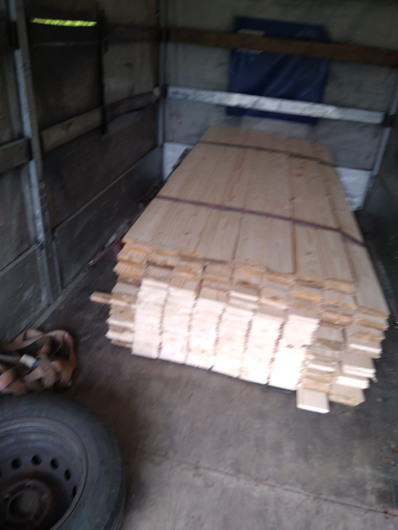 Stos desek zabezpieczony pasem w przestrzeni ładunkowej pojazdu, widoczne opony i pasy transportowe.