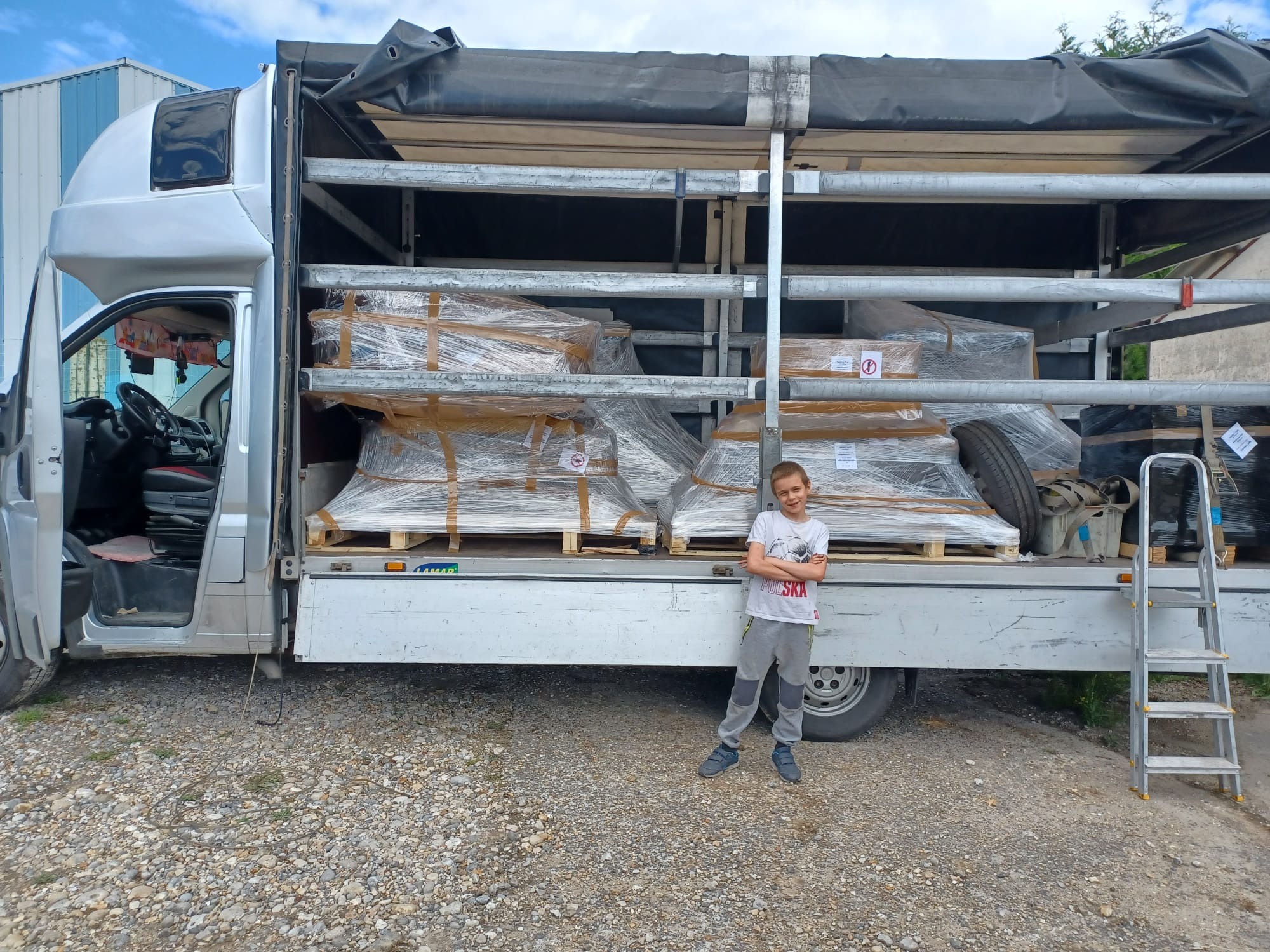 Chłopiec stoi przed otwartą furgonetką wypełnioną paletami z zapakowanymi elementami, na zewnątrz drabina, żwirowe podłoże.