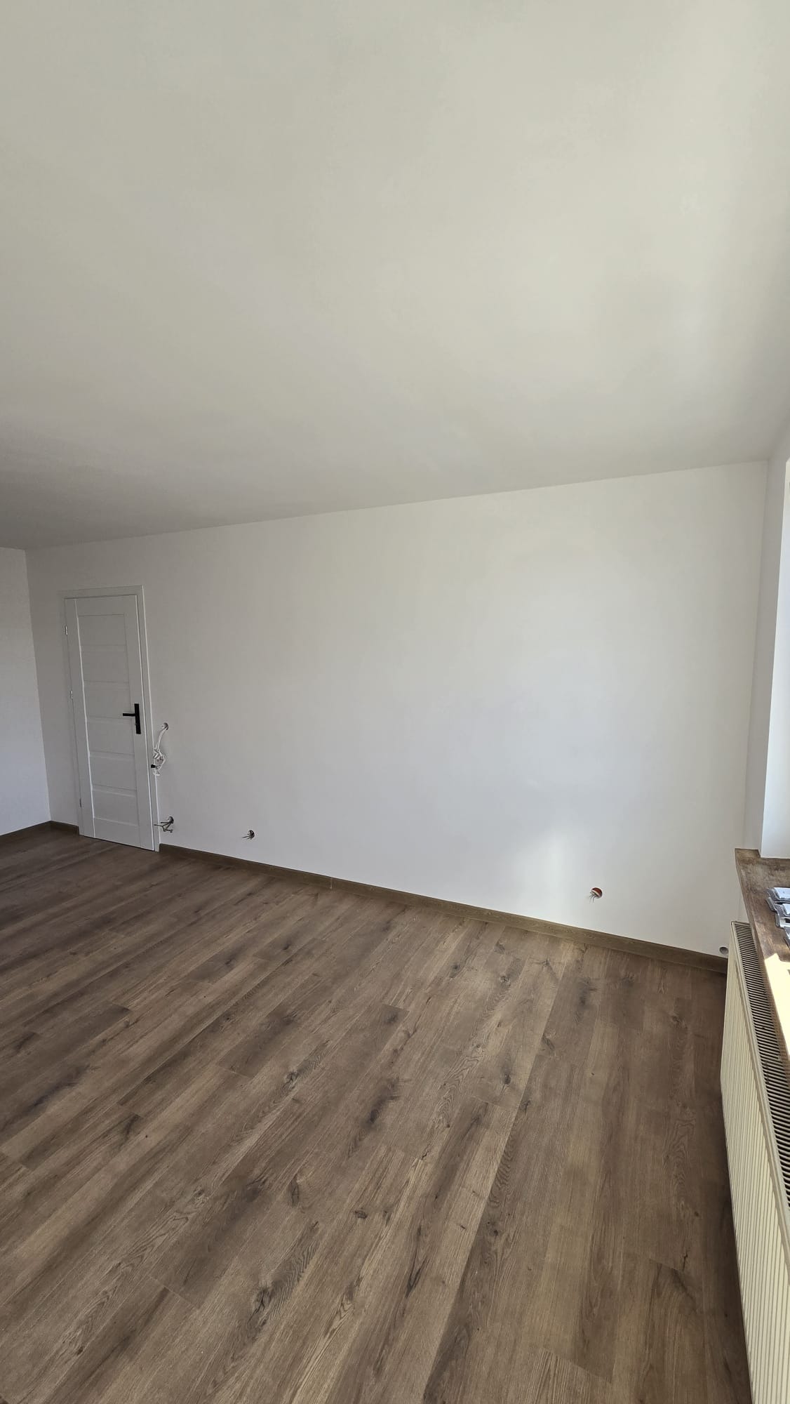 Jasne, przestronne wnętrze po remoncie z panelami podłogowymi imitującymi drewno i świeżo pomalowanymi na biało ścianami, widoczne wyprowadzenia elektryczne i grzejnik.