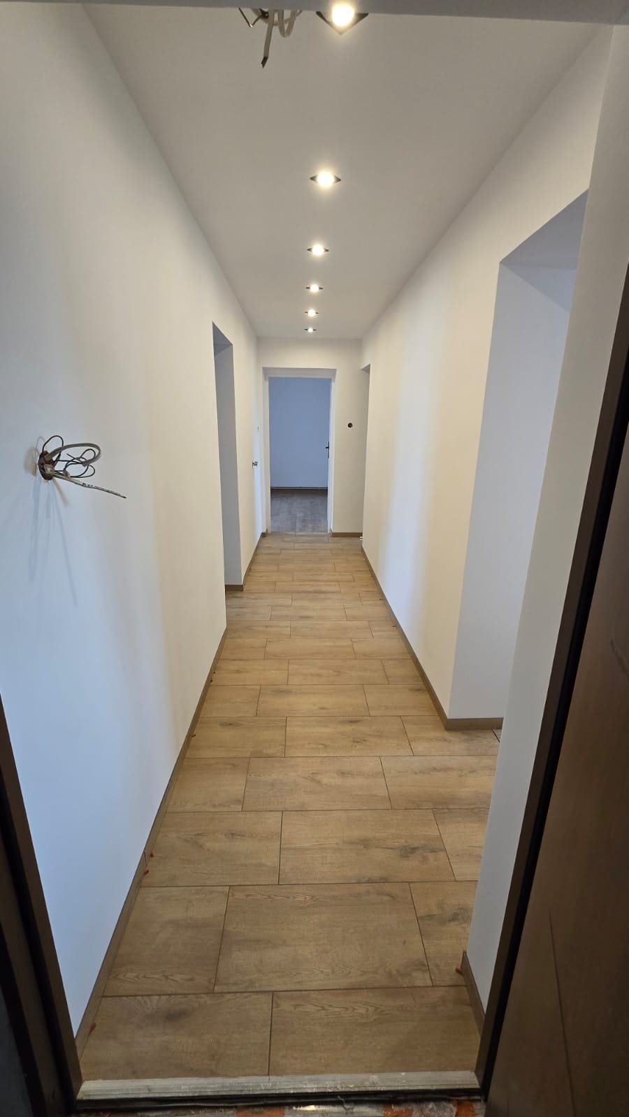 Długi korytarz z wykończonymi białymi ścianami, drewnopodobną podłogą z płytek i rzędem wpuszczonych punktowych lamp sufitowych; widoczne niedokończone okablowanie elektryczne.