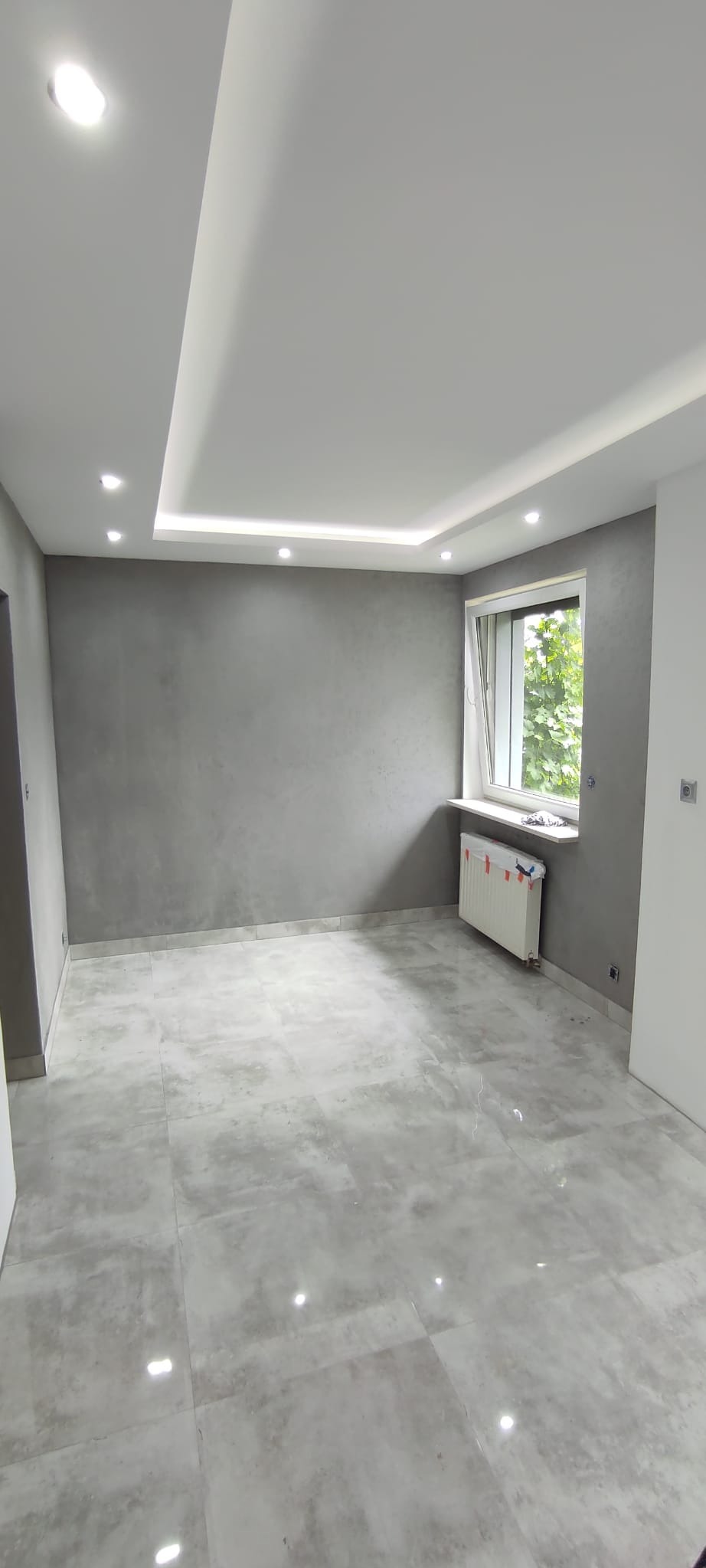 Pusty pokój po remoncie z szarymi ścianami z efektem dekoracyjnym, szarą podłogą z płytek, białym sufitem podwieszanym z oświetleniem punktowym i grzejnikiem pod oknem.