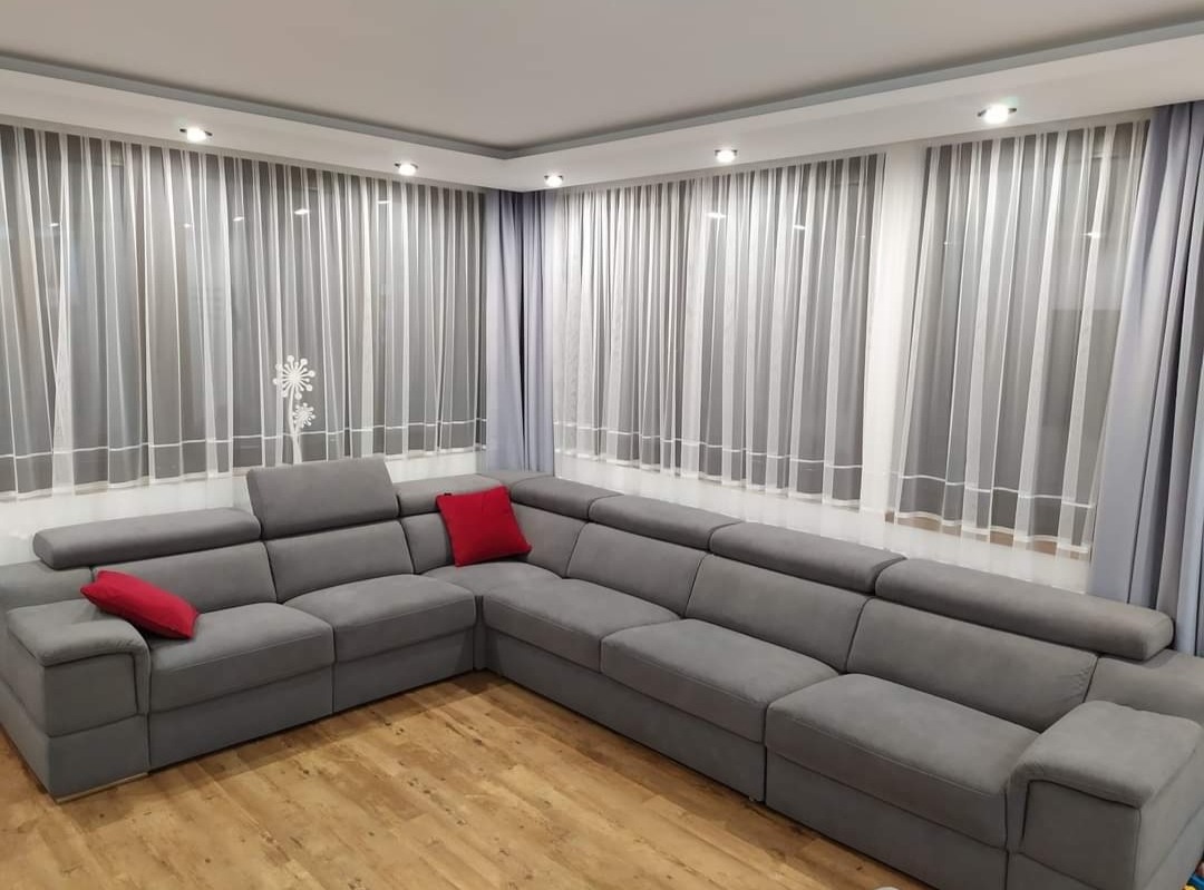 Jasne wnętrze salonu z dużym, szarym narożnikiem z regulowanymi zagłówkami i czerwonymi poduszkami, drewnianą podłogą i oknami zasłoniętymi białymi firanami z delikatnym wzorem.