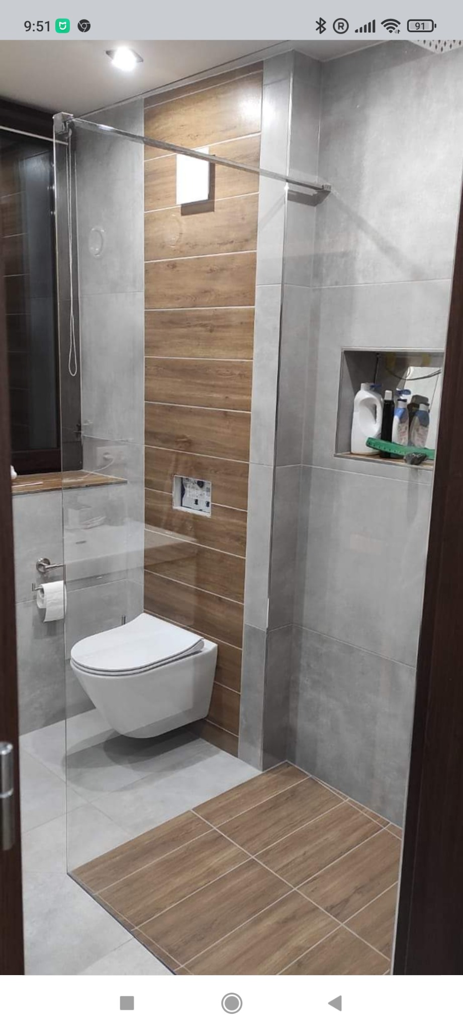 Nowoczesna łazienka z wiszącą toaletą, szklaną kabiną prysznicową, płytkami imitującymi drewno na ścianie i podłodze oraz wnęką z kosmetykami.