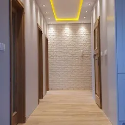 Długi korytarz z jasną podłogą z paneli, ściany pomalowane na biało, z trzema parami drewnianych drzwi po lewej stronie i zabudową po prawej, ściana na końcu korytarza wyłożona dekoracyjną cegłą...