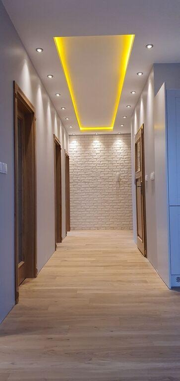 Długi korytarz z jasną podłogą z paneli, ściany pomalowane na biało, z trzema parami drewnianych drzwi po lewej stronie i zabudową po prawej, ściana na końcu korytarza wyłożona dekoracyjną cegłą...