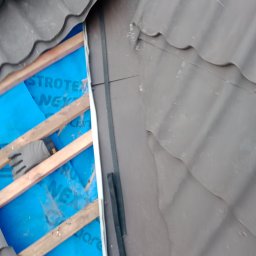 Art dach - Fragment dachu w trakcie naprawy: widoczne brązowe dachówki, drewniane łaty, niebieska membrana dachowa z nadrukiem, rękawica robocza.