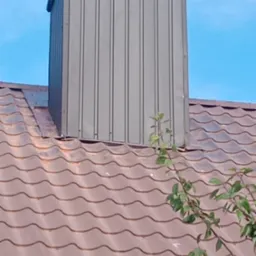 Szary komin wentylacyjny z blachy trapezowej na brązowym dachu z dachówki ceramicznej, widok z dołu, fragment gałęzi z liśćmi w prawym dolnym rogu.