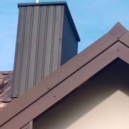Art dach - Szczegół dachu z brązowymi obróbkami blacharskimi i kominem z blachy trapezowej na tle błękitnego nieba.