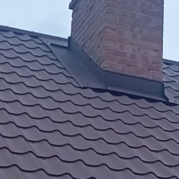 Art dach - Ciemnobrązowa blachodachówka z ceglanym kominem i metalowym wkładem kominowym na tle pochmurnego nieba.