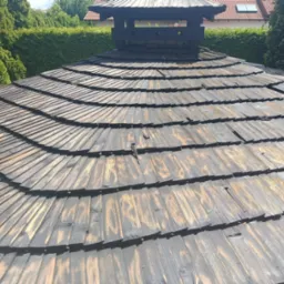 Unikalny, drewniany dach altany z kominem na tle zieleni. Widoczne detale konstrukcji i faktura drewna. Dach po renowacji, z elementami dekoracyjnymi.