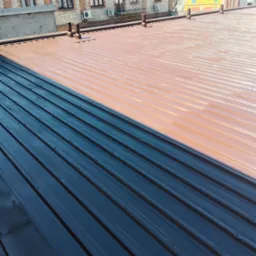 Dach z blachy trapezowej, częściowo pomalowany na ciemny kolor, z widocznymi kominami wentylacyjnymi i budynkami w tle.