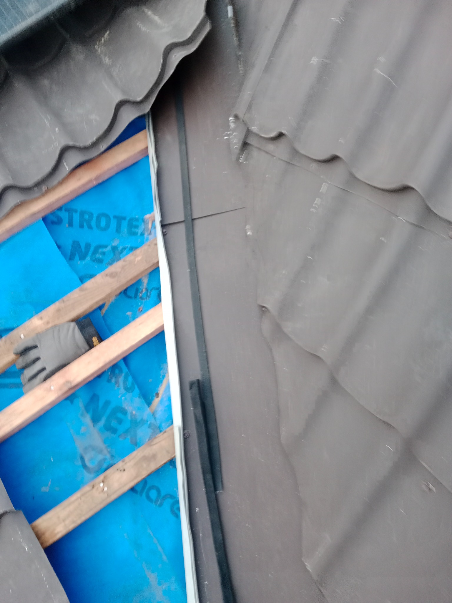 Fragment dachu w trakcie naprawy: widoczne brązowe dachówki, drewniane łaty, niebieska membrana dachowa z nadrukiem, rękawica robocza.