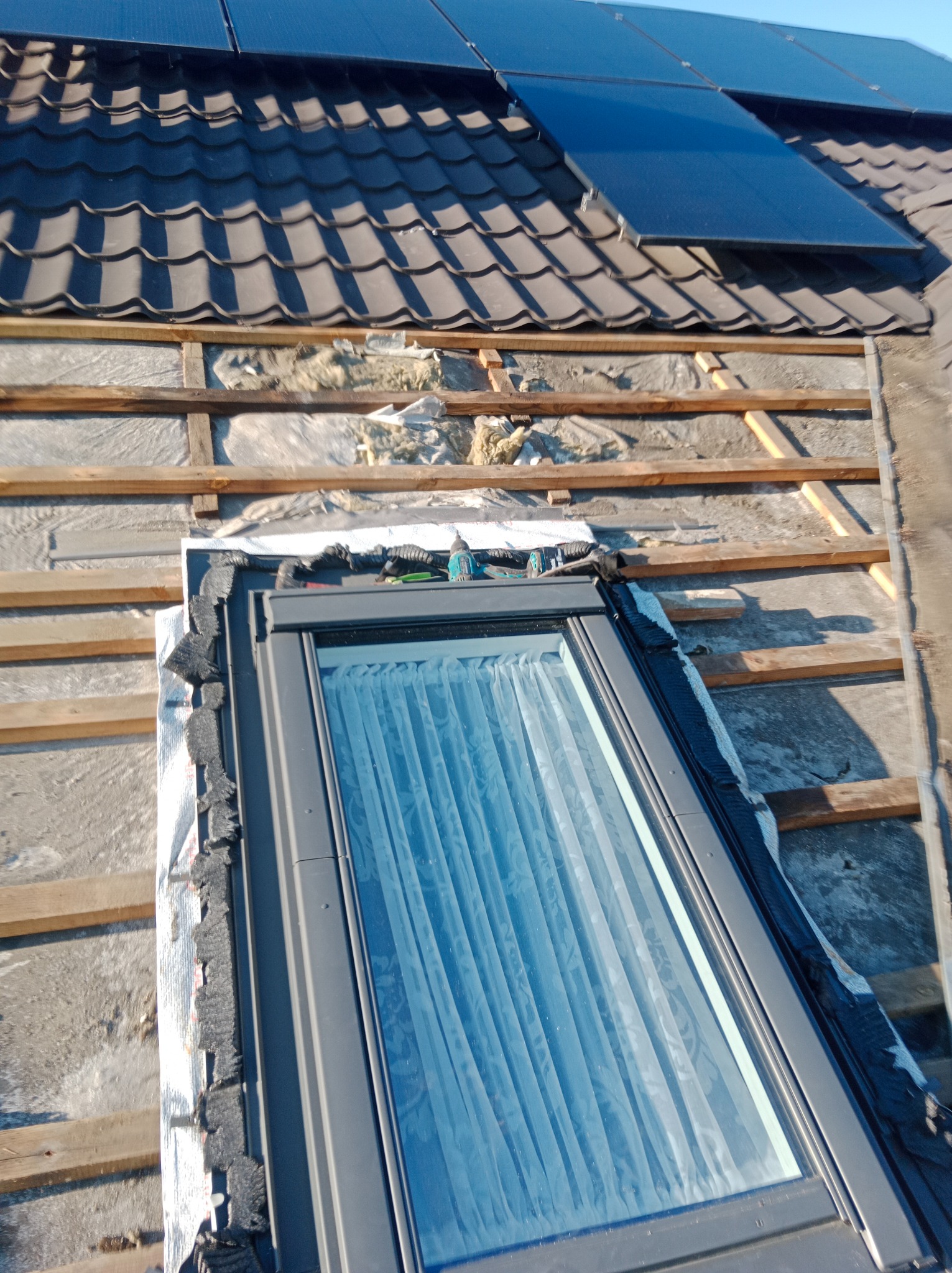 Montaż okna dachowego z zasłoniętą szybą, widoczne panele słoneczne na dachu z dachówką bitumiczną i elementy konstrukcji dachu