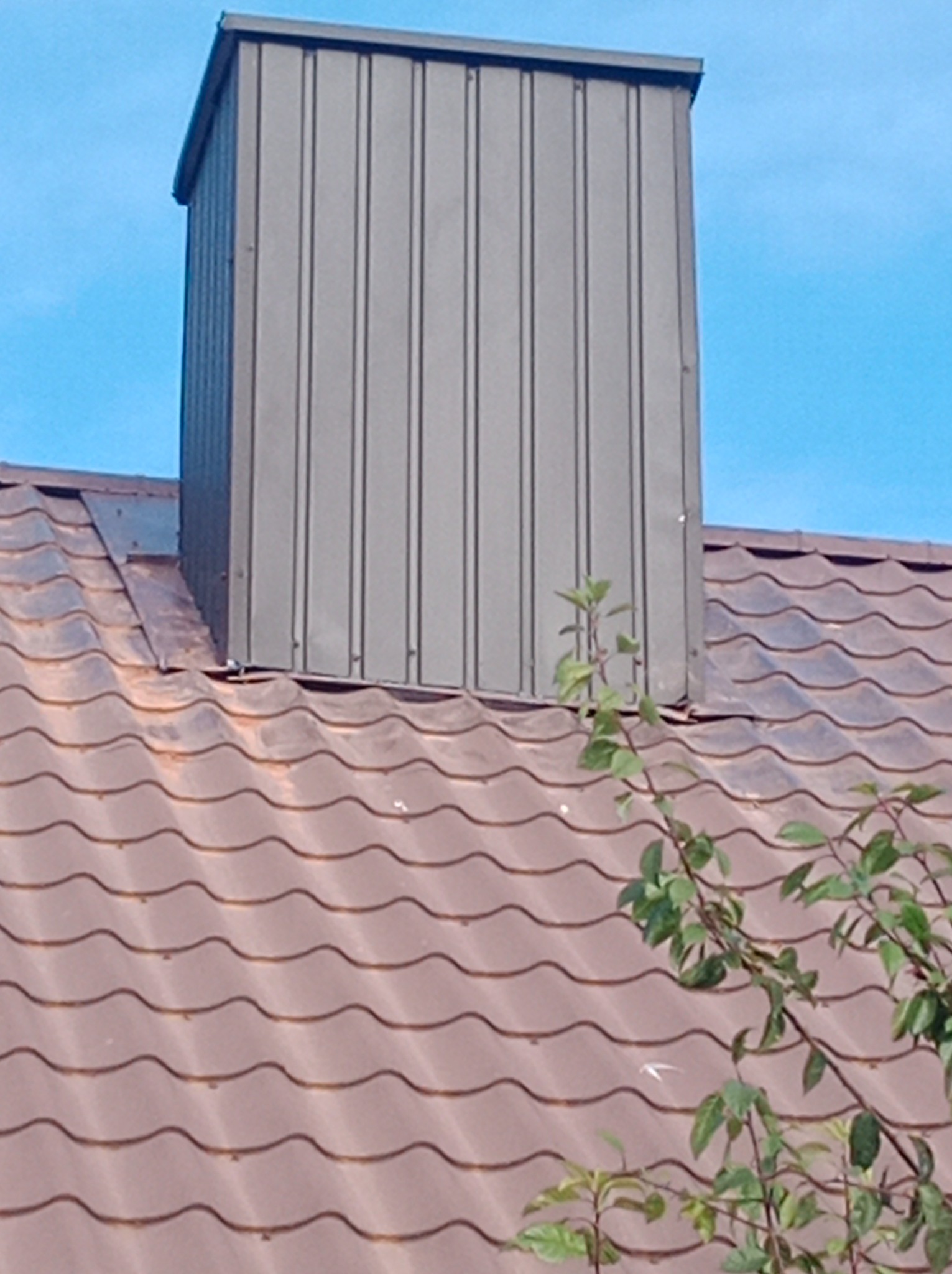 Szary komin wentylacyjny z blachy trapezowej na brązowym dachu z dachówki ceramicznej, widok z dołu, fragment gałęzi z liśćmi w prawym dolnym rogu.