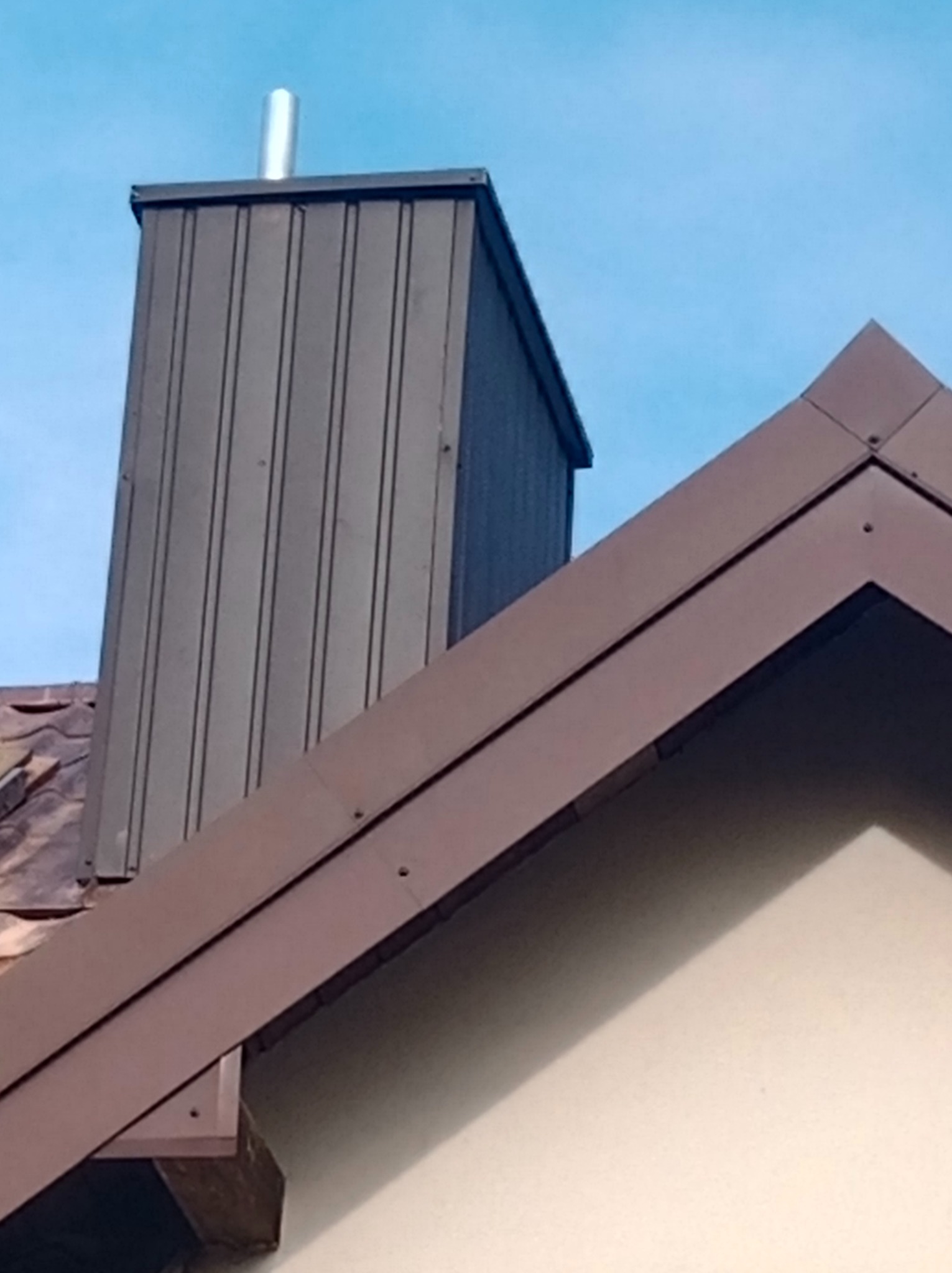 Szczegół dachu z brązowymi obróbkami blacharskimi i kominem z blachy trapezowej na tle błękitnego nieba.