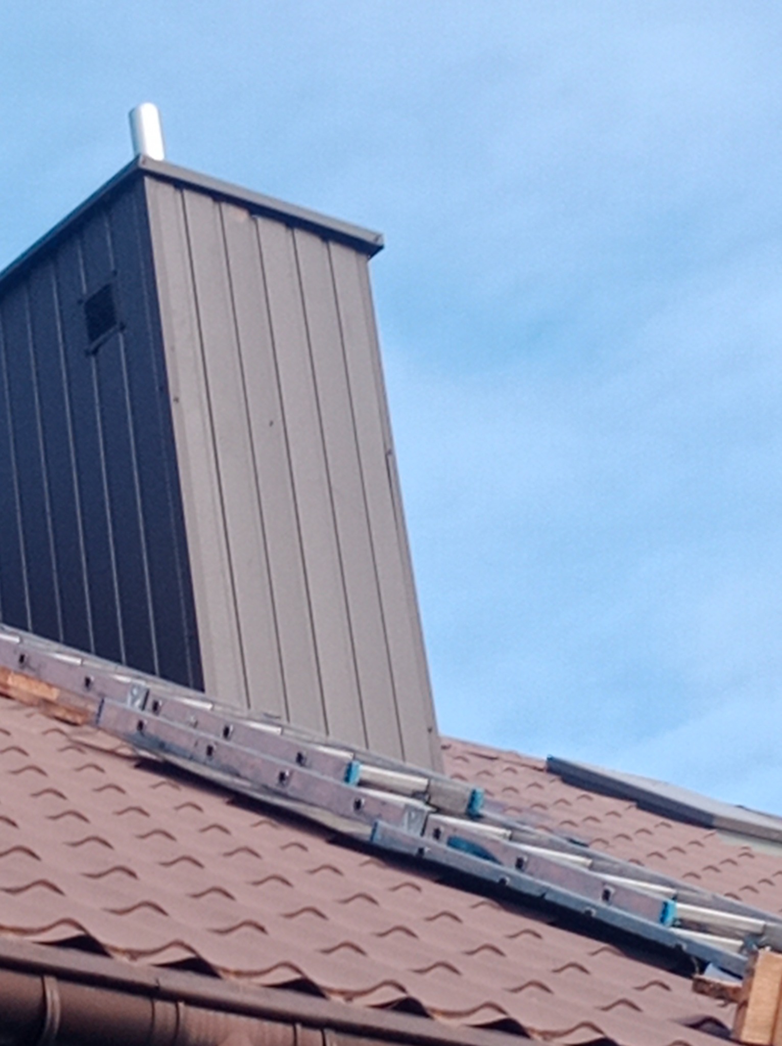 Szary komin wentylacyjny z blachy na brązowym dachu w Częstochowie, z rozłożoną drabiną opartą o jego podstawę, na tle błękitnego nieba.
