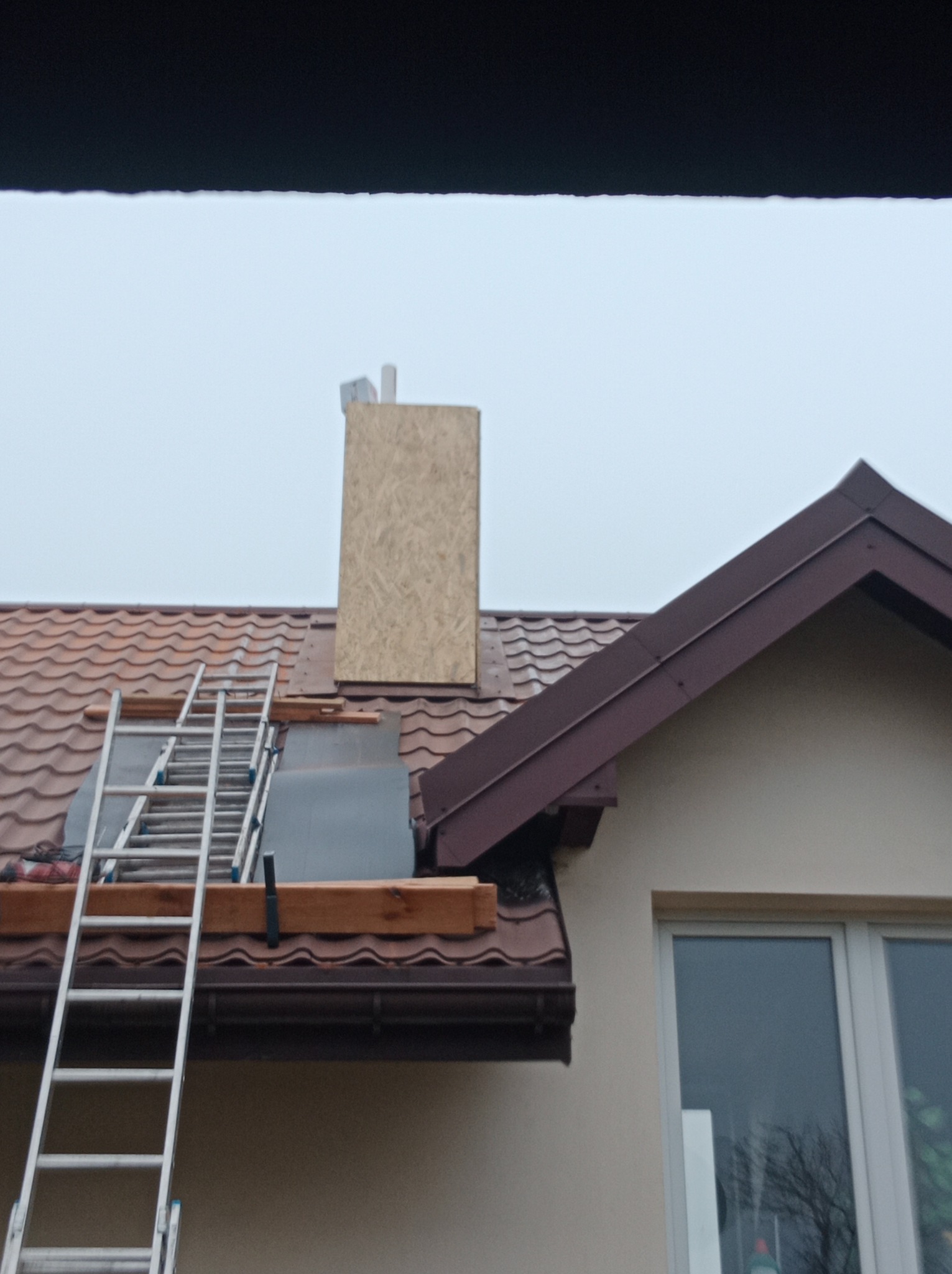 Dom z brązowym dachem podczas prac dekarskich. Widoczna drabina oparta o dach, drewniane elementy konstrukcyjne oraz obudowa komina z płyty OSB.