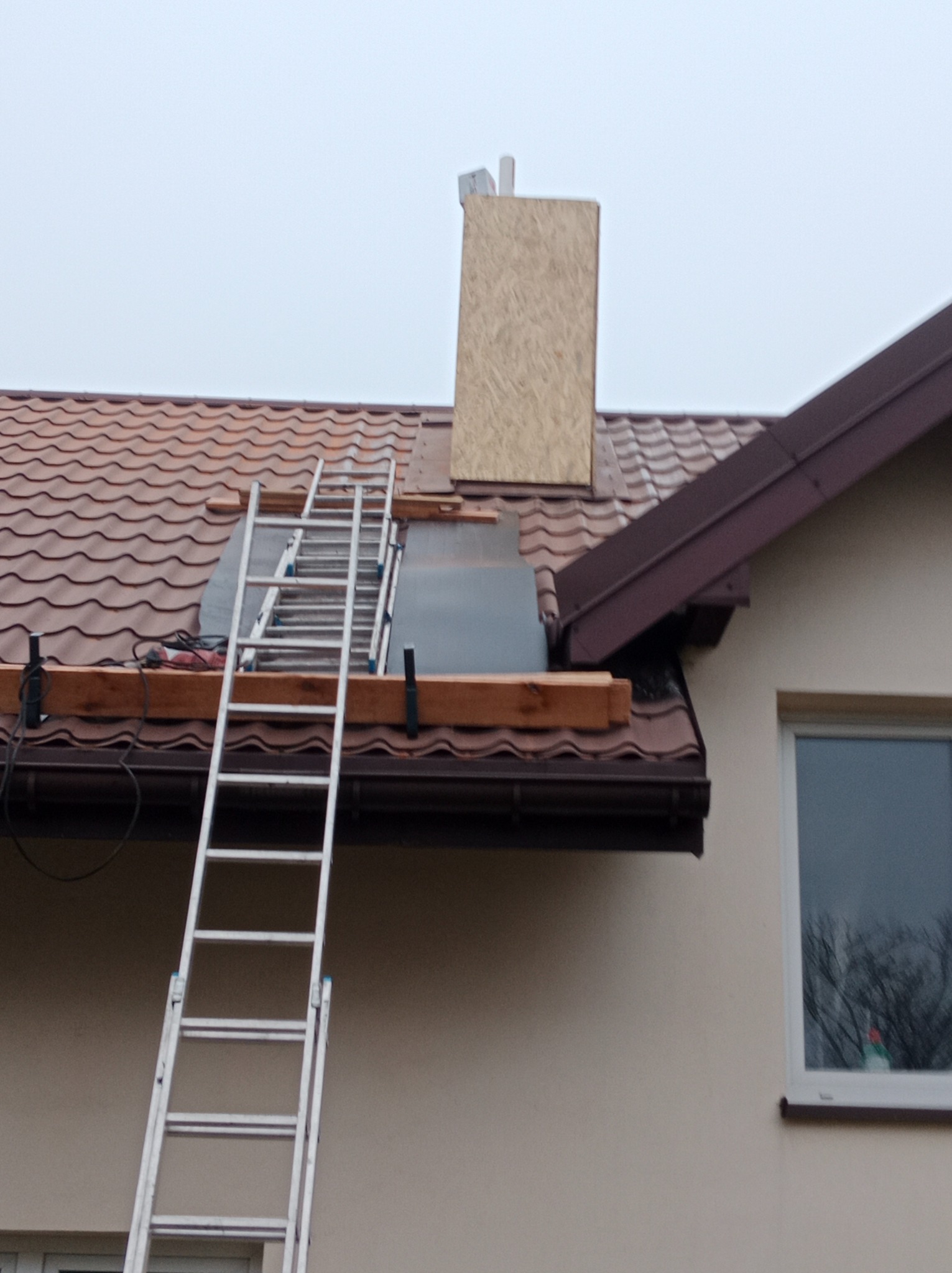 Praca na dachu: drabina oparta o brązowy dachówkowy dach, widoczne elementy konstrukcyjne komina obłożone płytą OSB, narzędzia na dachu, pochmurne niebo.