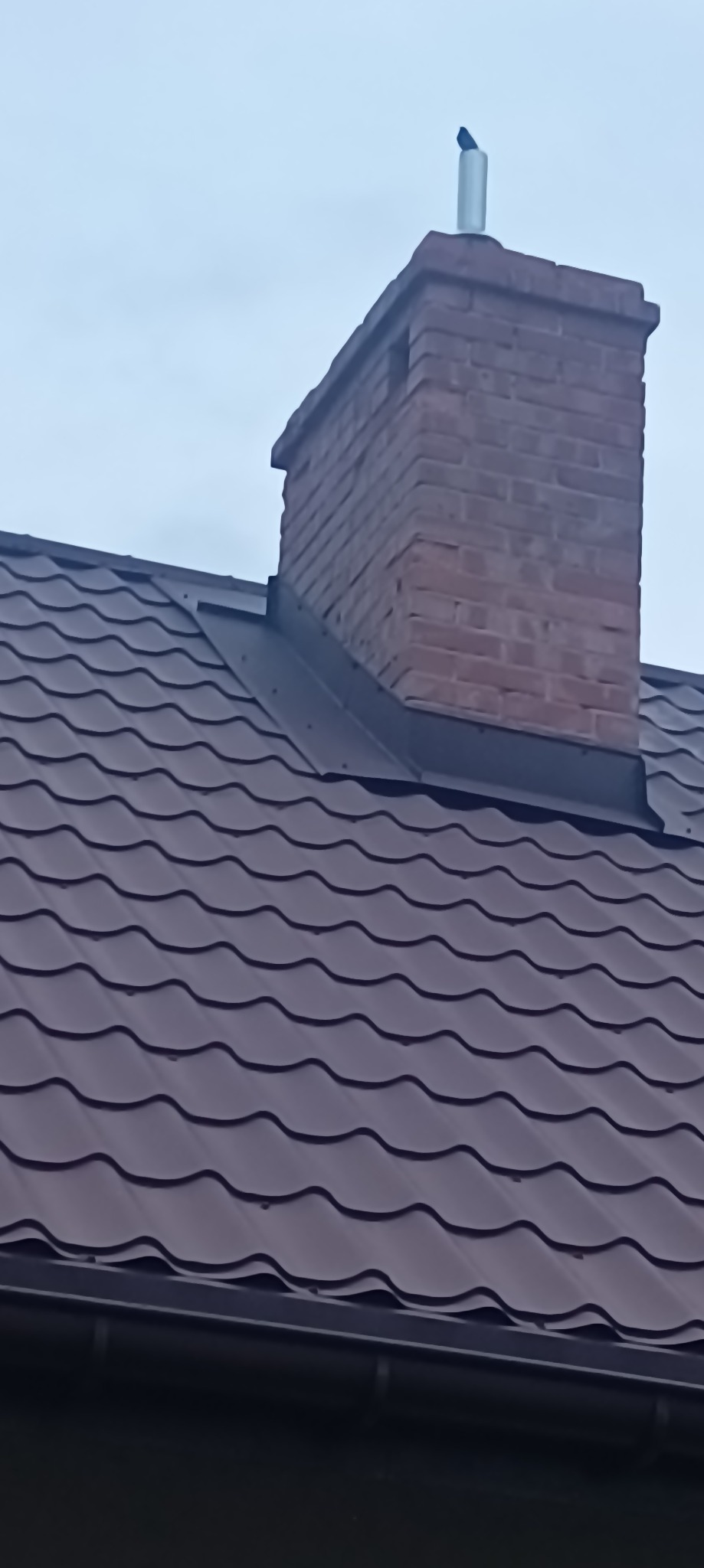 Ciemnobrązowa blachodachówka z ceglanym kominem i metalowym wkładem kominowym na tle pochmurnego nieba.