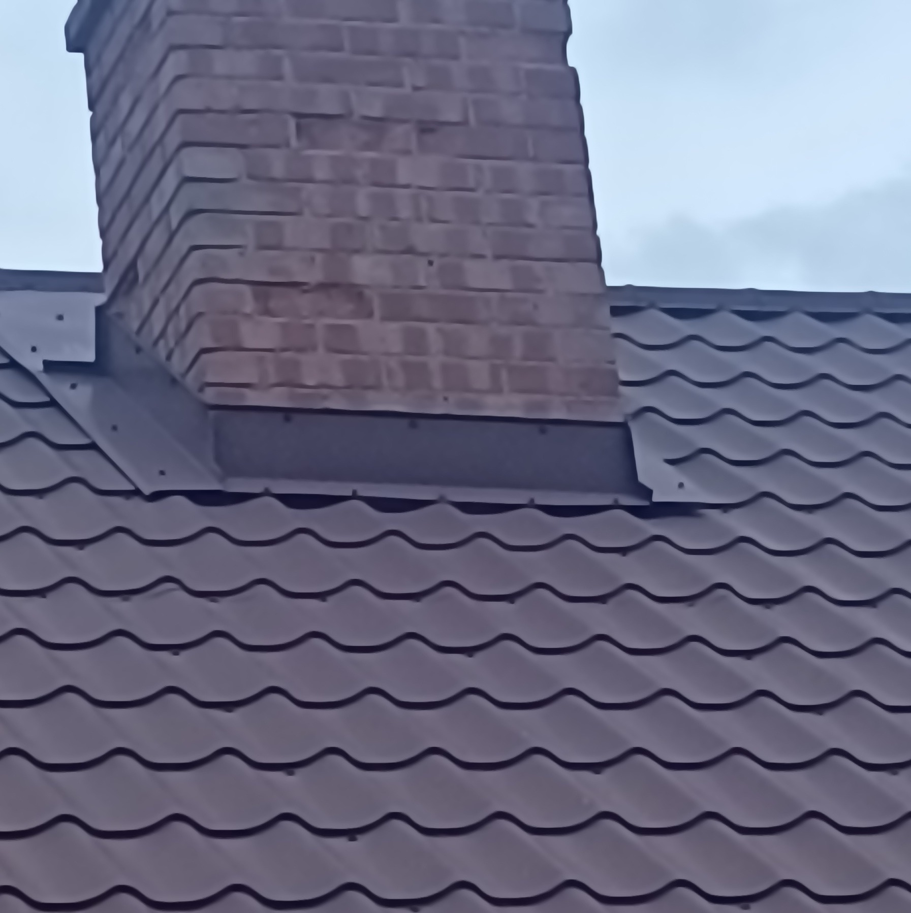 Brązowa blacha dachowa z ceglanym kominem, obróbka blacharska wokół komina, pochmurne niebo w tle.