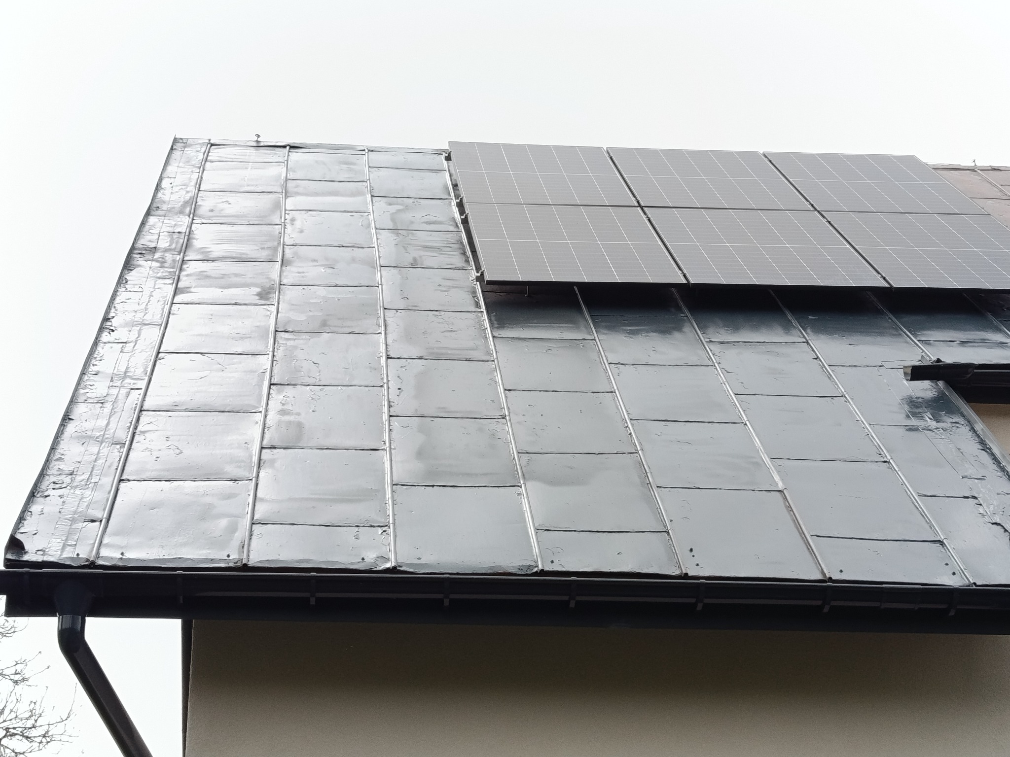 Dach pokryty metalową blachą w kształcie karo, z zamontowanymi panelami słonecznymi. Widoczny fragment orynnowania. Ujęcie z dołu, pochmurne niebo w tle.