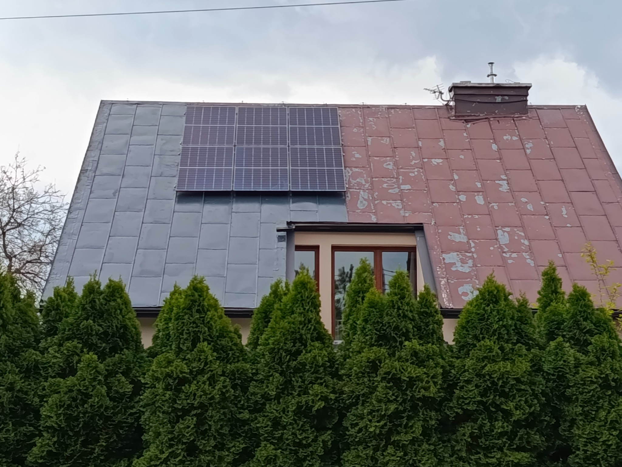 Dwuspadowy dach z panelami słonecznymi i kominem. Połowa dachu odnowiona, druga z łuszczącą się farbą. W tle zielone tuje i szare niebo.