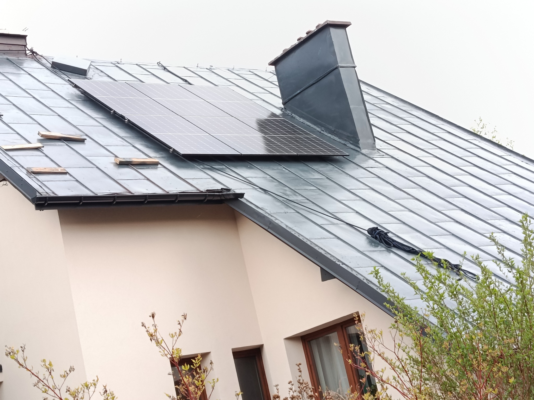 Metalowy dach z panelami solarnymi i kominem. Widoczne deski na dachu i lina. Dach w kolorze grafitowym. Fragment budynku w tle.