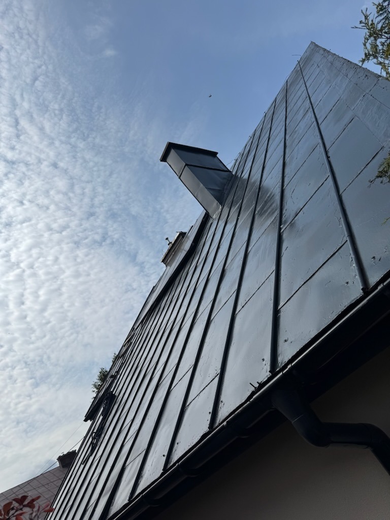 Błyszczący, ciemny dach z blachy trapezowej, widziany z dołu pod kątem. Widoczne rynny i fragment komina na tle błękitnego, zachmurzonego nieba.