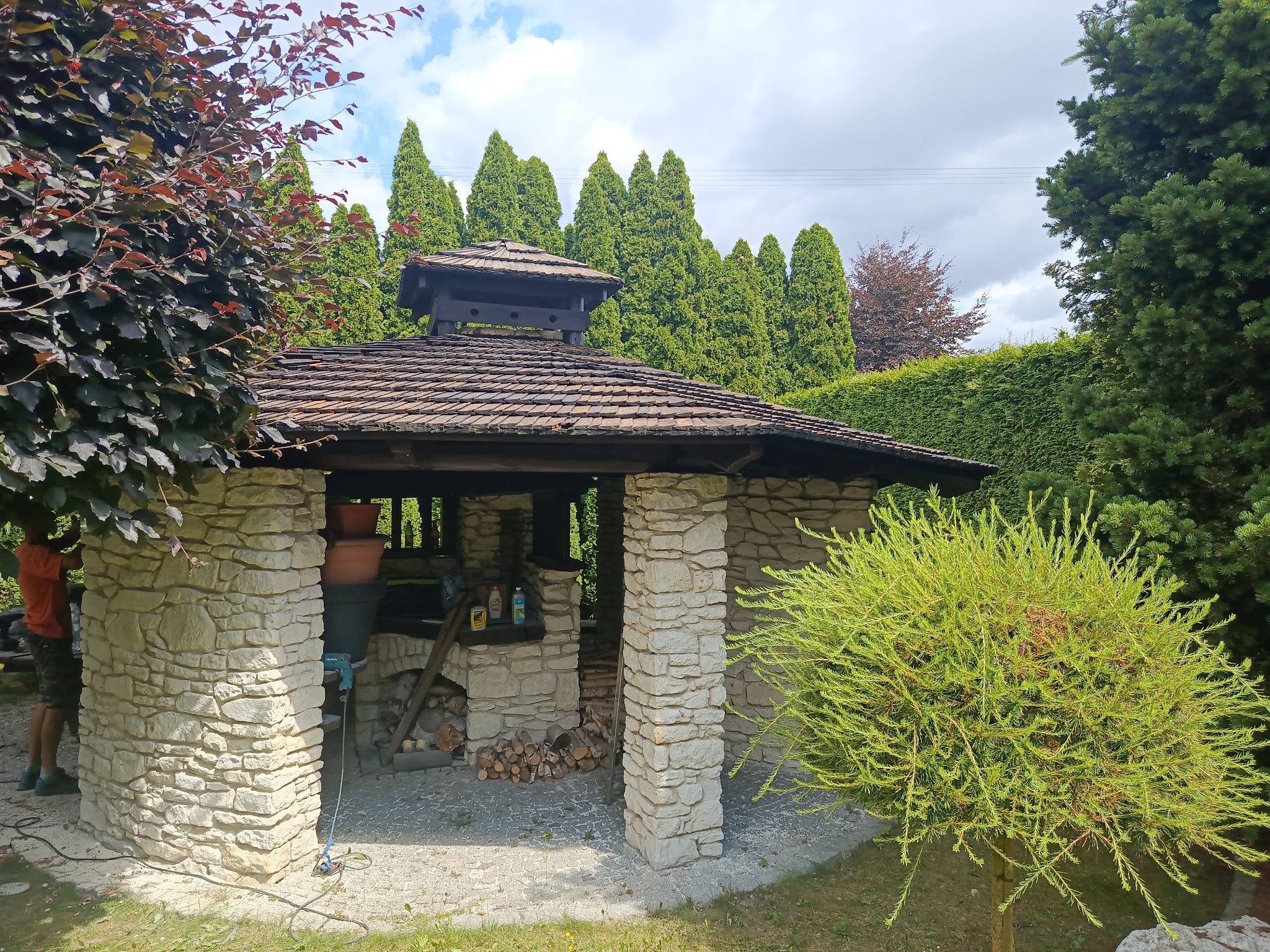 Kamienna altana z drewnianym dachem w ogrodzie, otoczona tujami. Na blacie wewnątrz altany widoczne narzędzia i preparaty. Osoba częściowo widoczna przy filarze.
