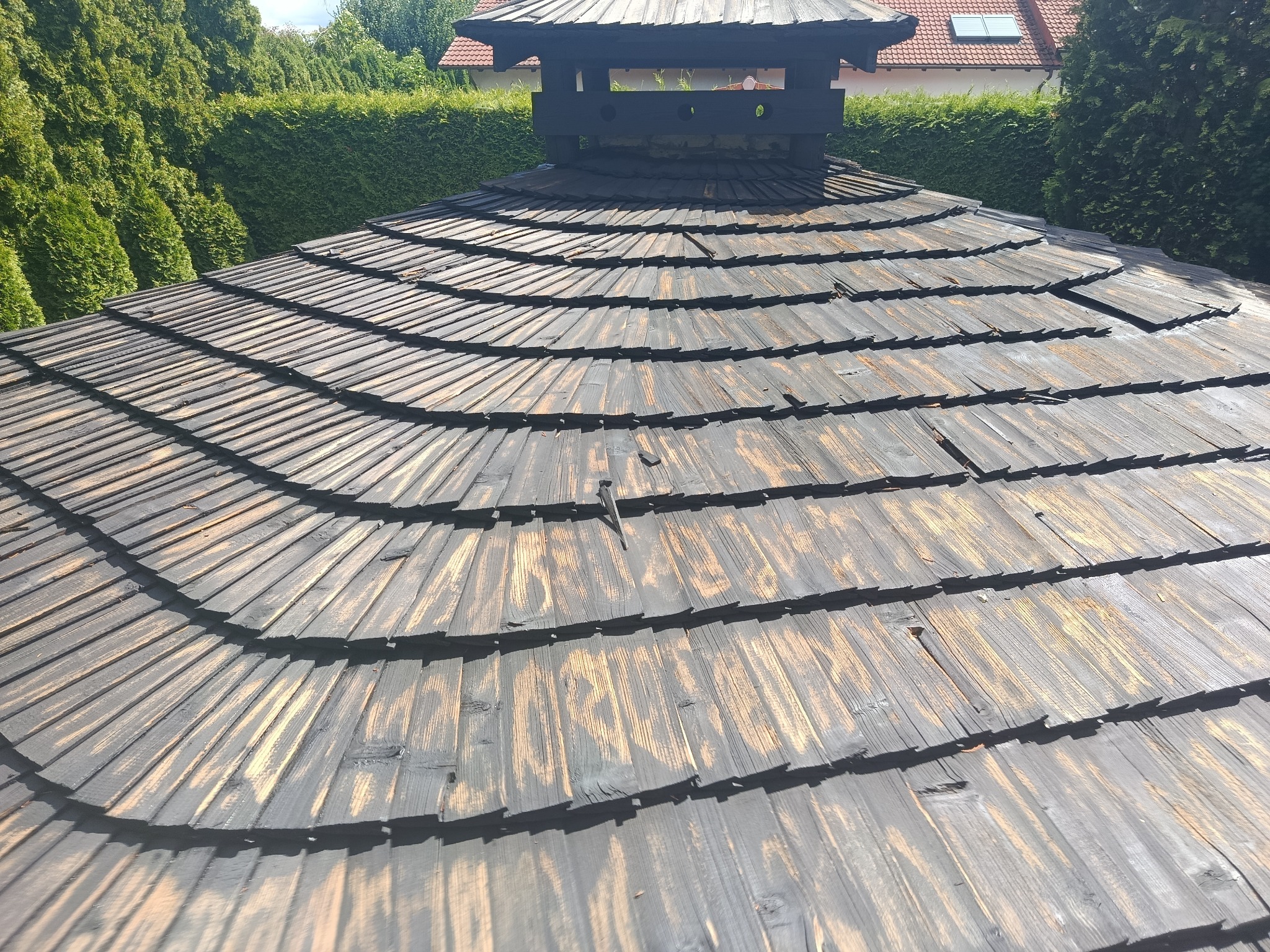 Unikalny, drewniany dach altany z kominem na tle zieleni. Widoczne detale konstrukcji i faktura drewna. Dach po renowacji, z elementami dekoracyjnymi.