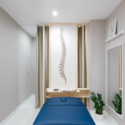Interfizjo S.A. - Jasne wnętrze gabinetu z niebieskim stołem do masażu, obrazem kręgosłupa w drewnianej ramie, lustrem i rośliną doniczkową.