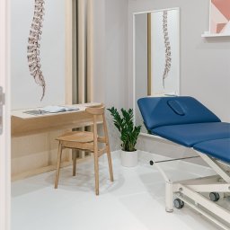 Interfizjo S.A. - Jasne wnętrze gabinetu z niebieskim leżanką rehabilitacyjną, biurkiem z książką, drewnianym krzesłem i rysunkiem kręgosłupa na ścianie oraz w lustrze.