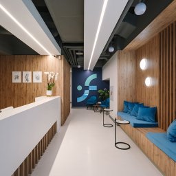 Interfizjo S.A. - Nowoczesne wnętrze kliniki w Warszawie z recepcją, drewnianymi panelami, błękitnymi akcentami i minimalistycznym oświetleniem.