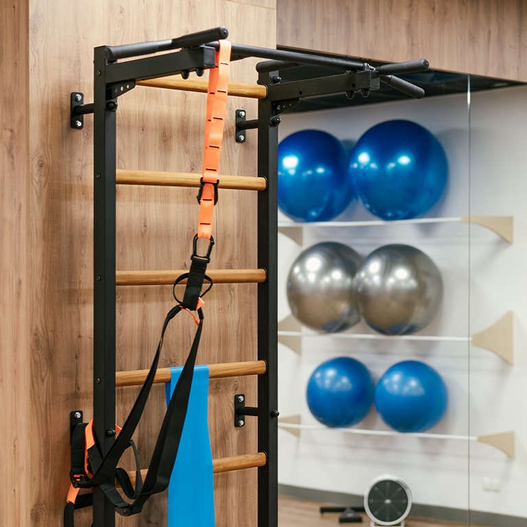Drabinka gimnastyczna z zawieszonymi taśmami TRX i gumą oporową, tło z piłkami do ćwiczeń na półkach, drewniana ściana w tle.