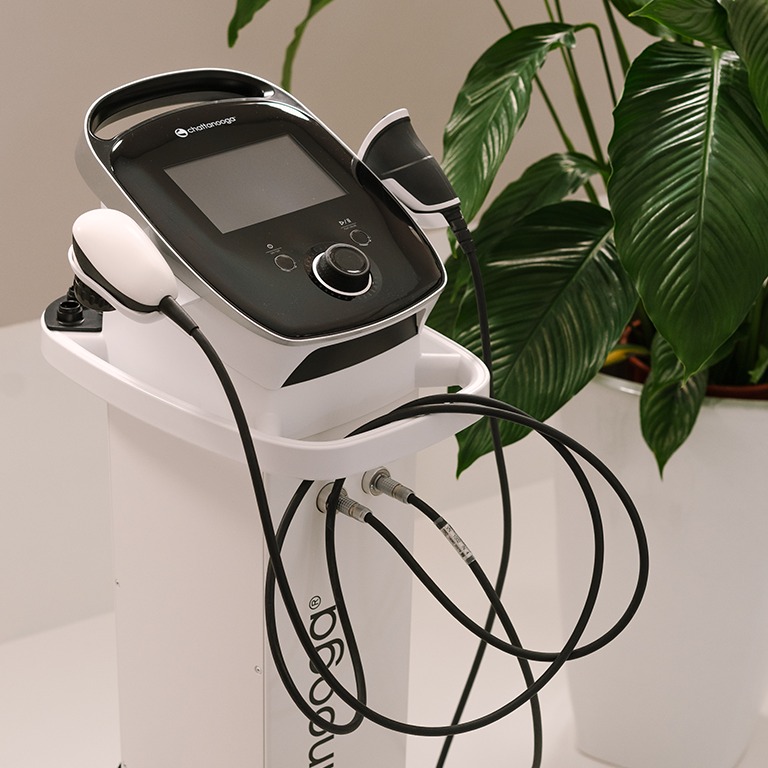 Aparat do fizykoterapii marki Chattanooga na białym wózku, z widocznymi głowicami i kablami, na tle zielonej rośliny doniczkowej.