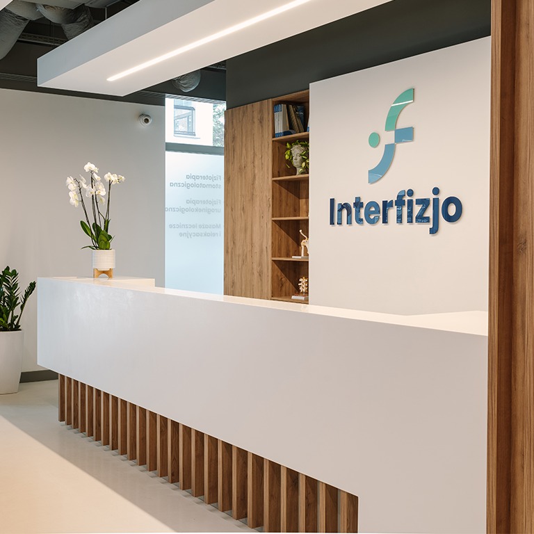 Nowoczesna recepcja kliniki z białym blatem, drewnianymi akcentami i logo 'Interfizjo' na ścianie, uzupełniona roślinami i regałem z książkami.