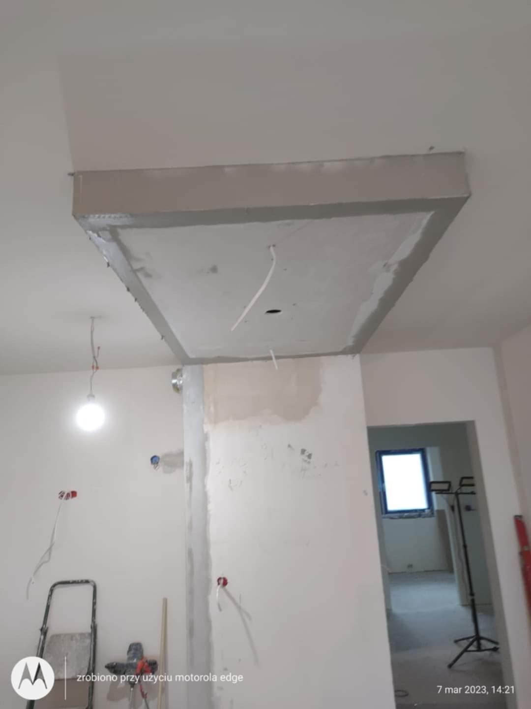 Sufit podwieszany w trakcie montażu: widoczne kable, surowe wykończenie, ściany w trakcie prac. W tle okno i drabina malarska.