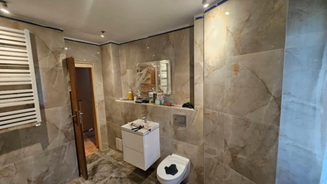 Wykończona łazienka z płytkami imitującymi marmur, białą szafką pod umywalką i lustrem. Widoczny grzejnik drabinkowy i częściowo otwarte drzwi.