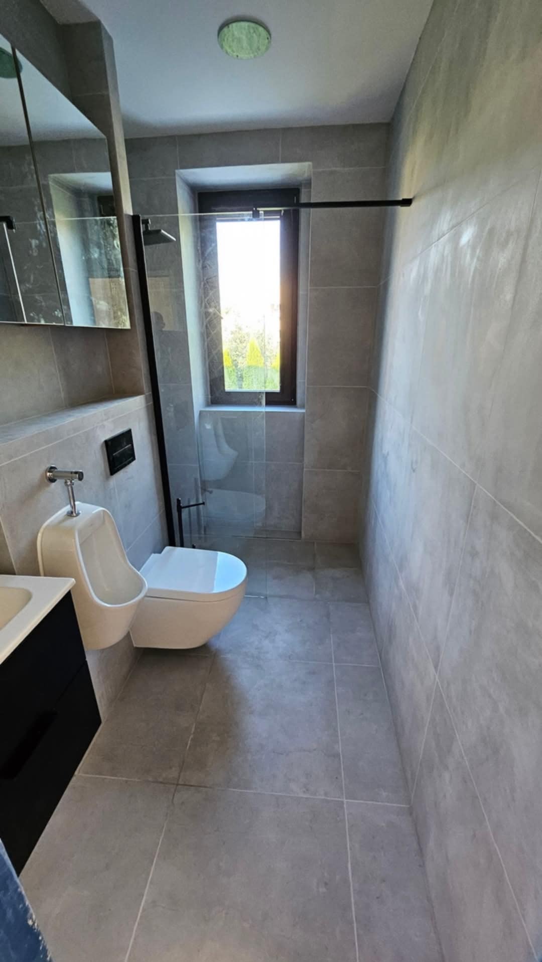 Szara, nowoczesna łazienka z kabiną prysznicową typu walk-in, toaletą i pisuarze. Okno z widokiem na zieleń. Minimalistyczny design, płytki w odcieniach szarości.