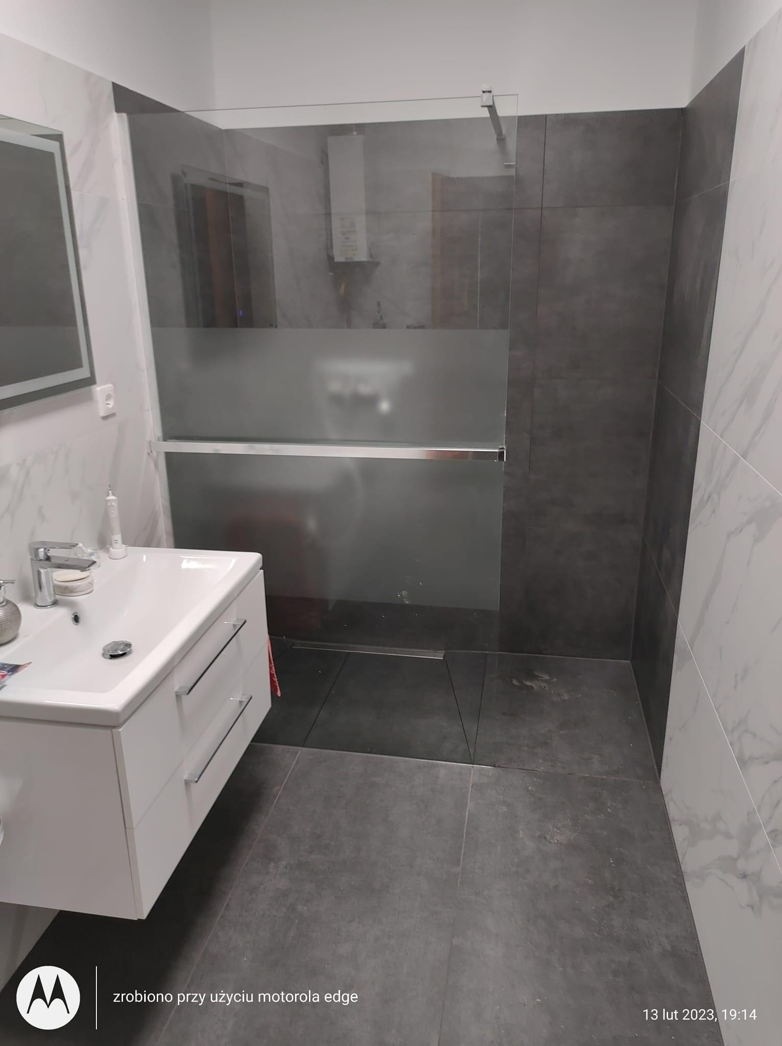 Nowoczesna łazienka z kabiną prysznicową typu walk-in z częściowo matową szybą, białą umywalką z szafką i szarą podłogą z płytek. Na ścianach połączenie białych płytek imitujących marmur i szarych...