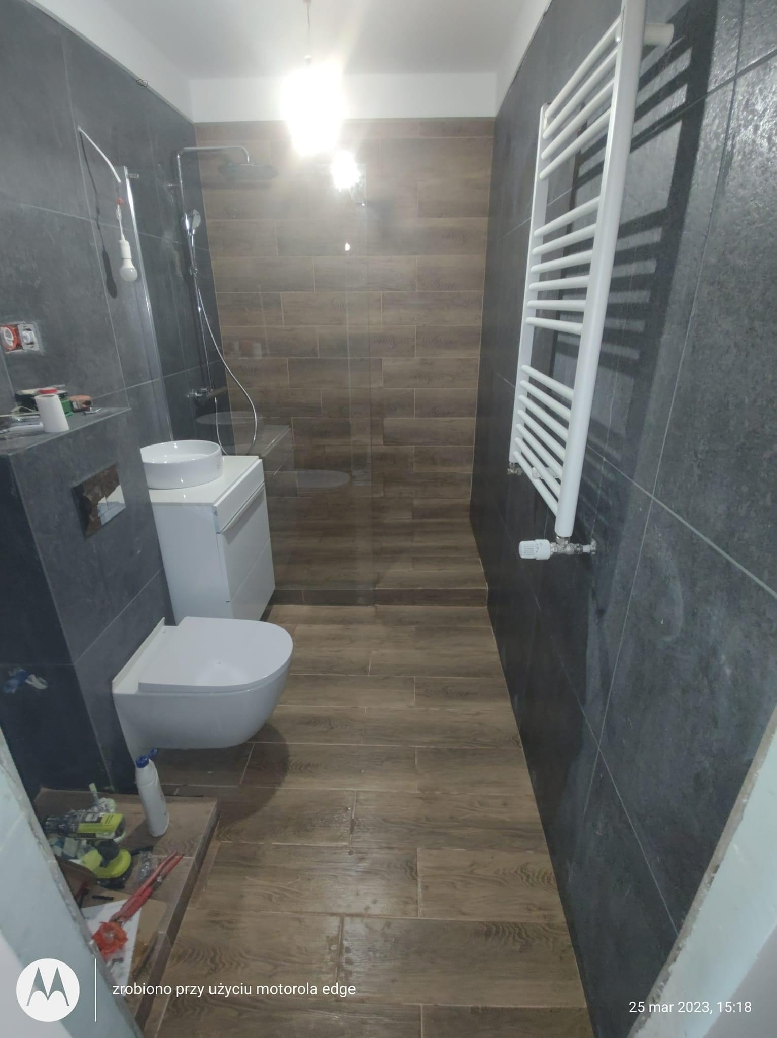 Nowoczesna łazienka w trakcie wykańczania: ciemne płytki na ścianach kontrastują z drewnopodobną podłogą, widoczny biały grzejnik drabinkowy, umywalka nablatowa na szafce i toaleta, narzędzia...