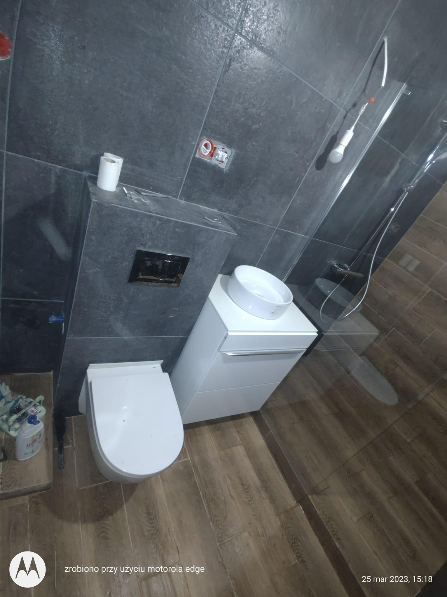 Nowoczesna łazienka w trakcie wykańczania: szare płytki na ścianach, drewnopodobne płytki na podłodze, biała umywalka nablatowa i toaleta podwieszana z czarnym przyciskiem spłukiwania, widoczny...