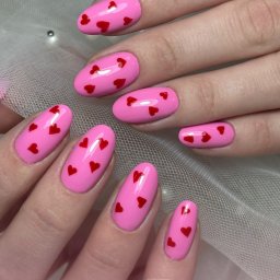 Pędzlem i pudrem - Różowe paznokcie w kształcie migdała z czerwonymi serduszkami, na tle delikatnej, białej tkaniny. Romantyczny, walentynkowy wzór na zadbanych dłoniach.