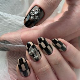 Pędzlem i pudrem - Dłoń z manicure w szachownicę beżowo-czarną, zdobiona srebrnymi krzyżami. Efektowny, błyszczący design na tle delikatnej, białej tkaniny.