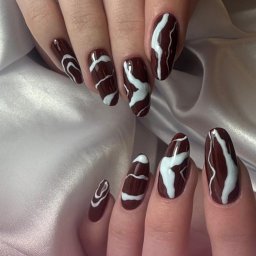 Pędzlem i pudrem - Dłonie z brązowymi paznokciami zdobionymi białymi, abstrakcyjnymi wzorami na satynowym tle. Widoczne logo 'PEDZLEM.I.PUDREM'.