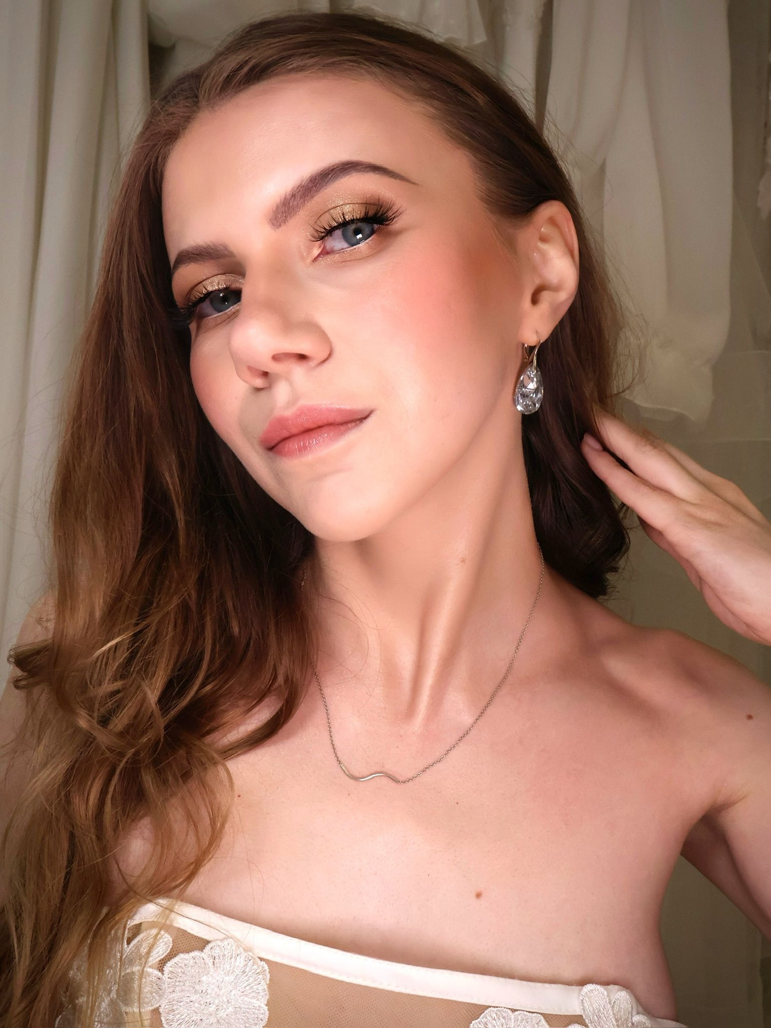 Portret kobiety z delikatnym makijażem, złotym cieniem na powiekach i błyszczącymi kolczykami, dłoń przy szyi, subtelna biżuteria, miękkie światło.