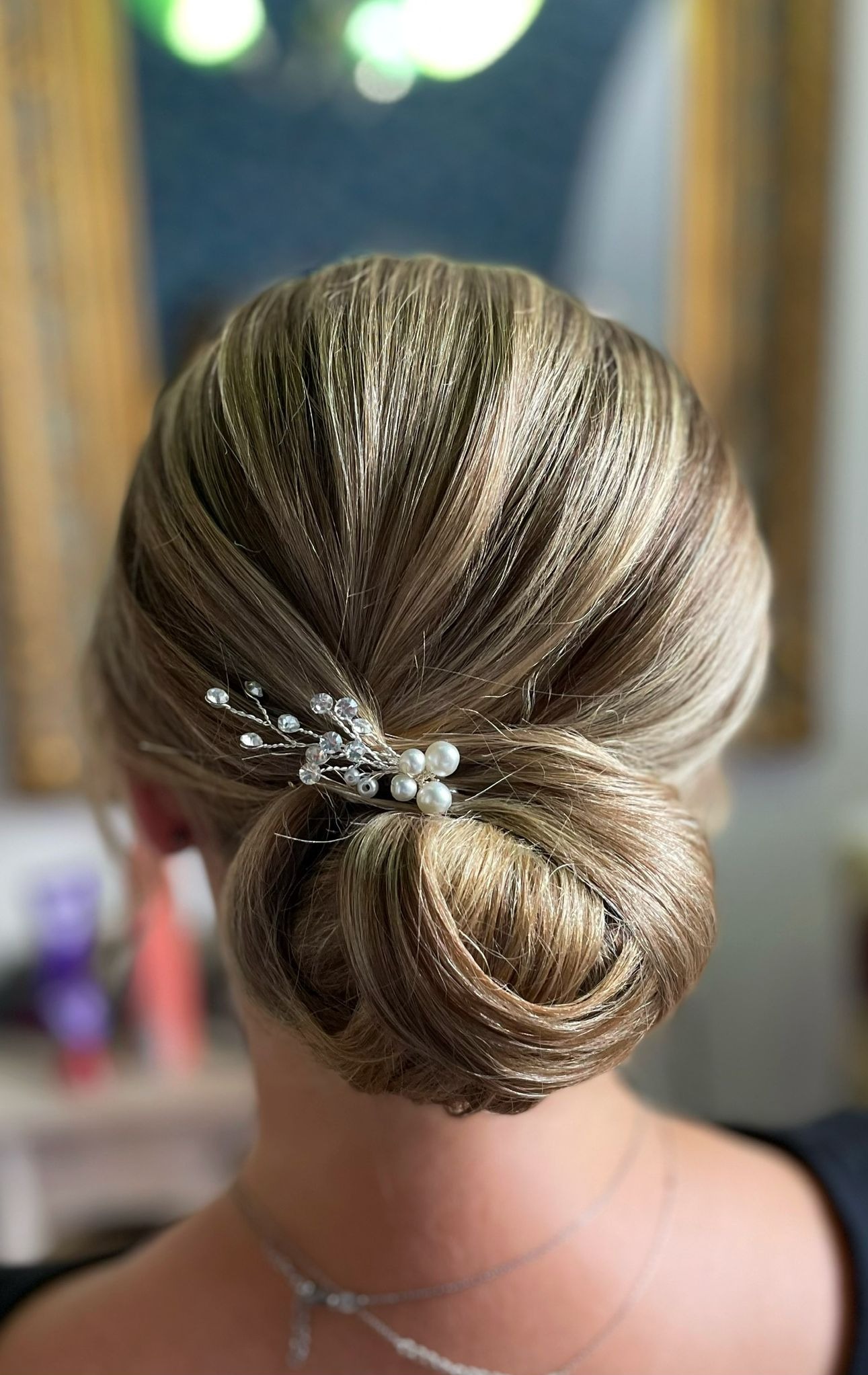 Eleganckie upięcie blond włosów z ozdobną spinką z pereł i kryształków, widok z tyłu, delikatny makijaż, idealne na specjalną okazję.