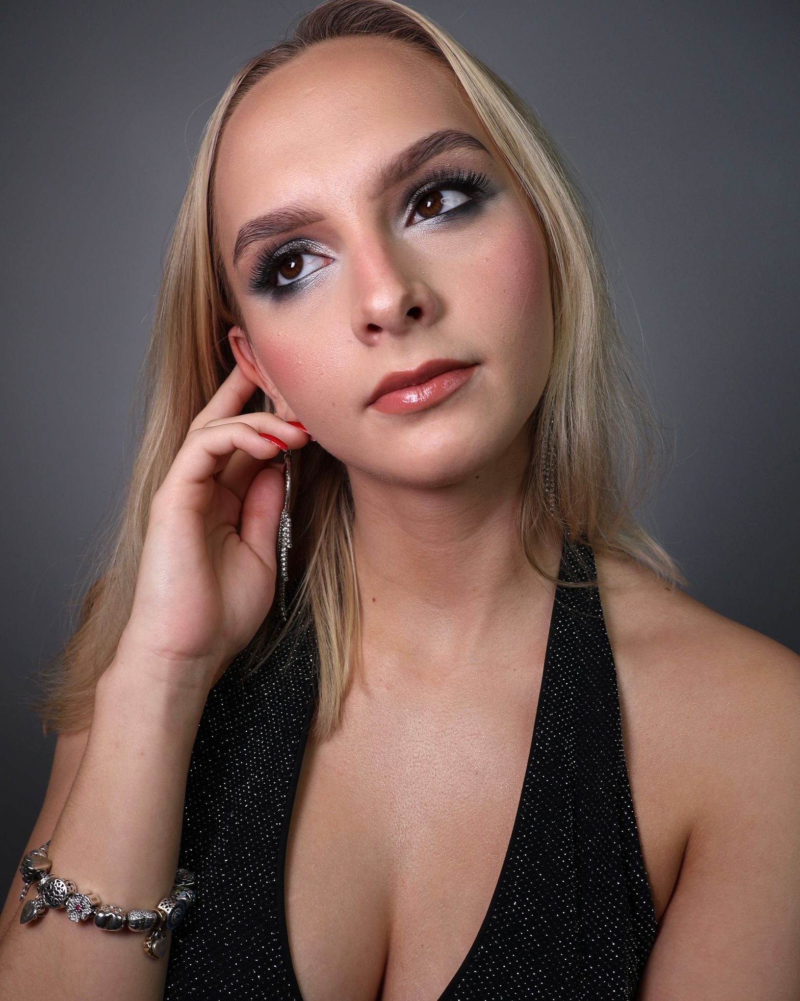 Portret kobiety z profesjonalnym makijażem oczu w odcieniach szarości i błyszczącymi ustami. Widoczna biżuteria: kolczyk i bransoletka. Ciemne tło.